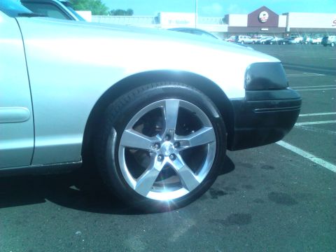 1999 Ford Crown Victoria - A silver car's front fender and wheel are visible, parked in a lot with a building in the background.
