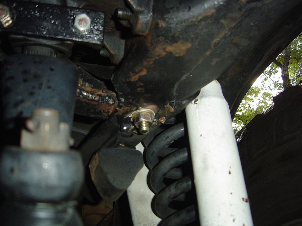 Close-up of a rusty steering box bracket with a bolt and nut, near a coil spring and white shock absorber