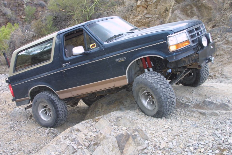 Blue and tan lifted Ford Bronco flexing over desert rocks, 3/4 front view with red shocks
