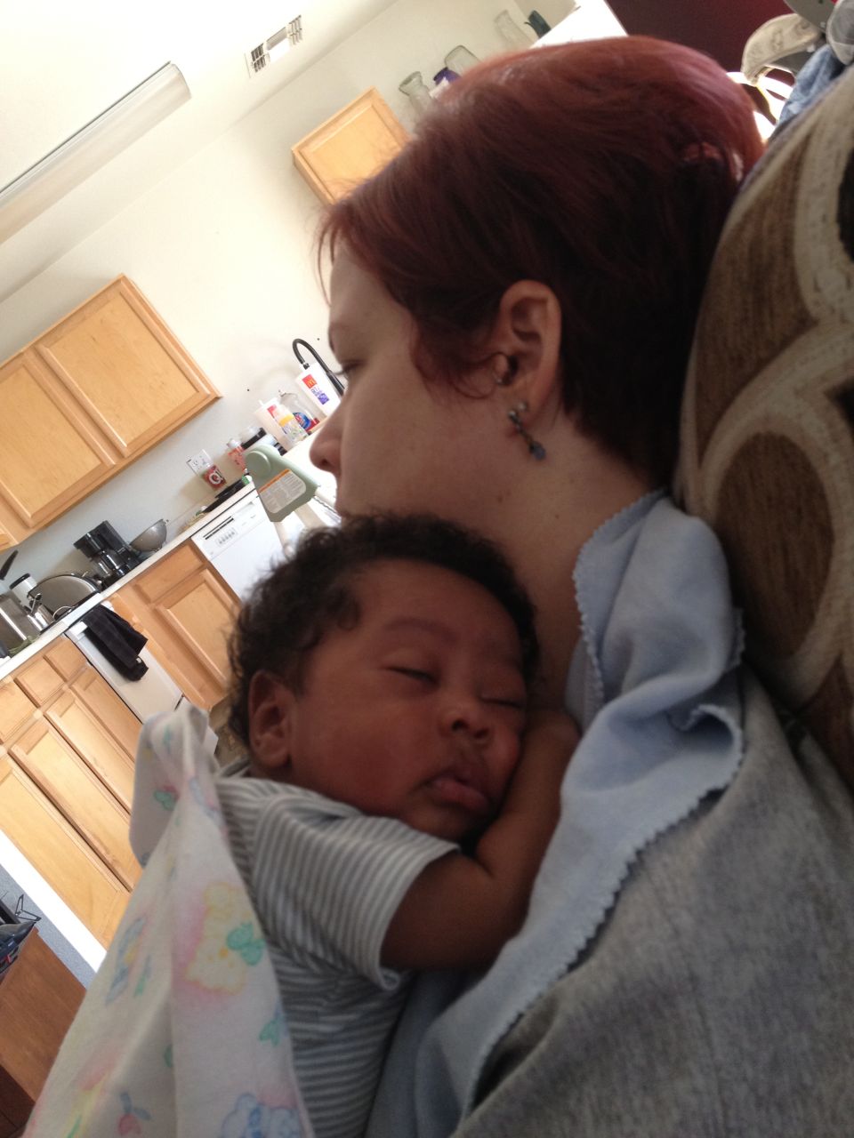 Woman with red hair holding a sleeping baby in her arms. Kitchen cabinets visible in background
