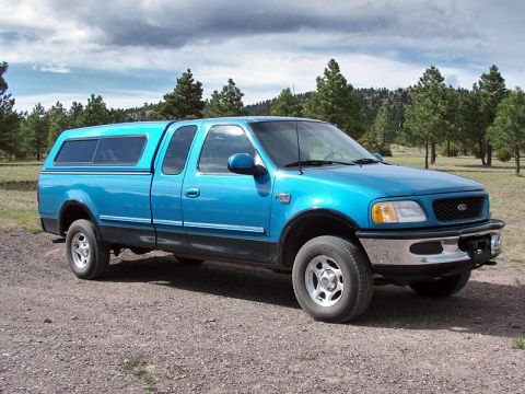 1998 Ford F-150 - Blue 1998 Ford F-150 XLT SuperCab pickup truck with a blue camper shell parked on a gravel surface.