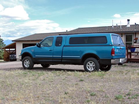1998 Ford F-150 - A blue pickup truck with a camper shell is parked in front of a house on a cloudy day.