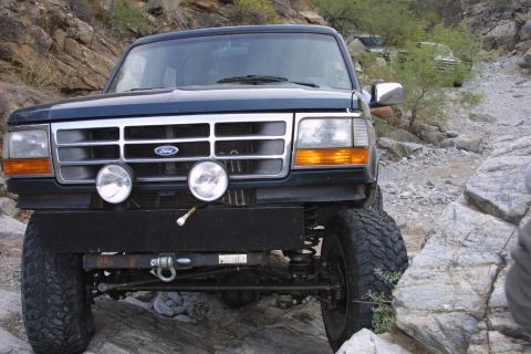 1994 Ford Bronco - A dark blue Ford Bronco navigates a rocky off-road path, with another vehicle visible in the background.