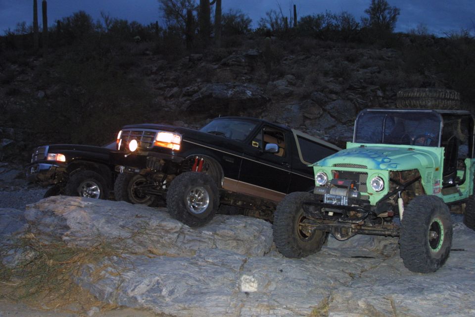 Lifted black and tan Ford Bronco on desert rocks at dusk beside a green Jeep