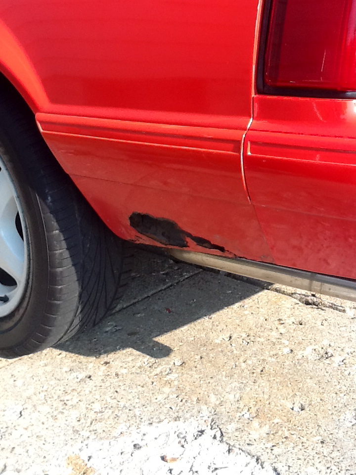 Red car side panel with rust near the wheel well and a chrome trim piece