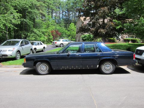 1991 Ford LTD Crown Victoria - A dark blue sedan is parked on the street in front of a house with other vehicles nearby.