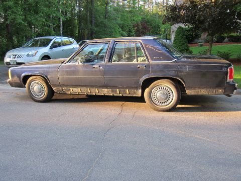 1991 Ford LTD Crown Victoria - A dark blue sedan is parked on a cracked asphalt road with another car behind it.