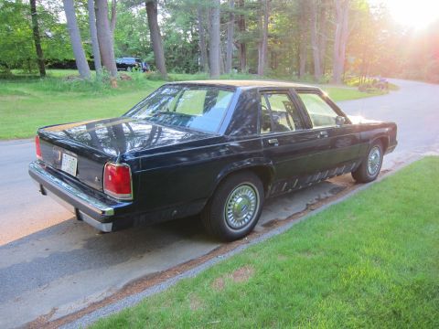 1991 Ford LTD Crown Victoria - A dark sedan is parked on the side of a road with trees and grass in the background.