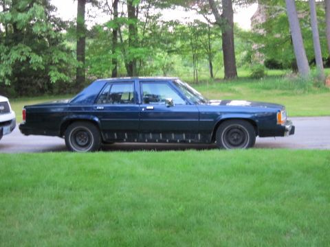 1991 Ford LTD Crown Victoria - Dark blue sedan with black lower body trim parked on a grassy area next to a road.