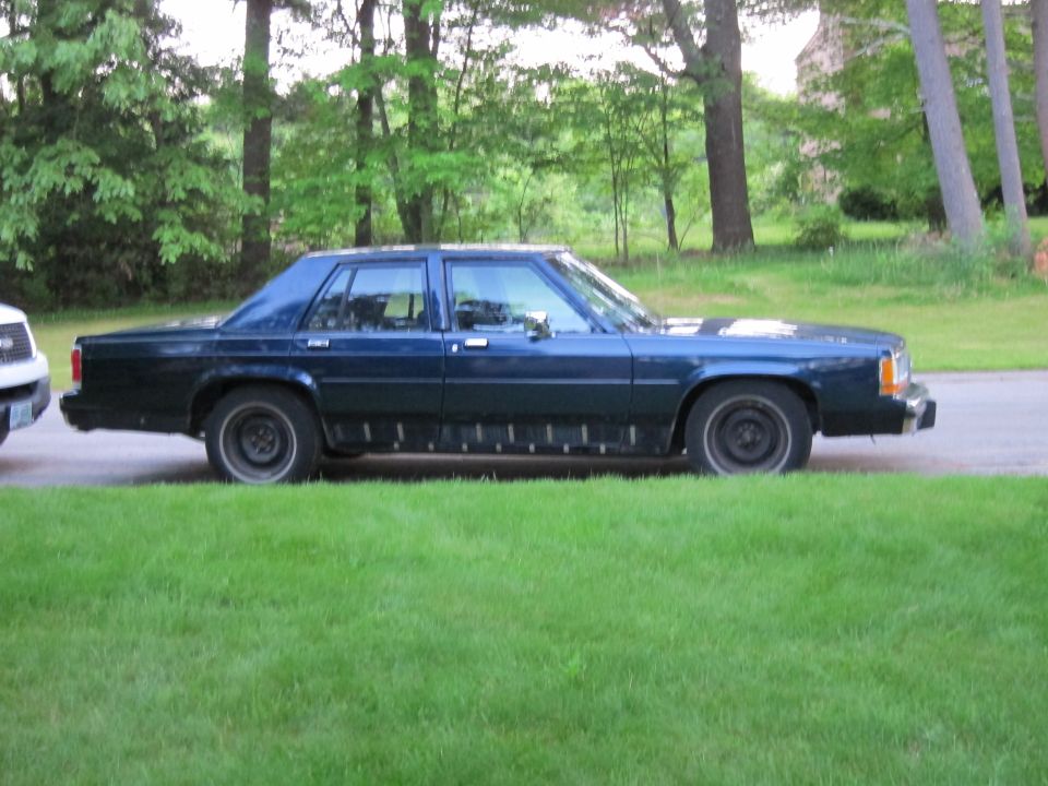 Side view of a dark blue sedan with black trim along the lower body panels, parked on grass next to a road. The front of a white vehicle is partially visible on the left