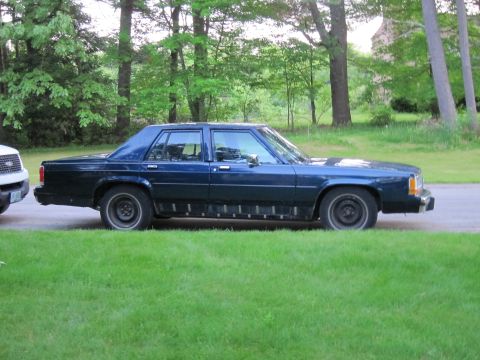 1991 Ford LTD Crown Victoria - A dark blue sedan is parked on the side of the road next to a white SUV, with trees and grass in the background.