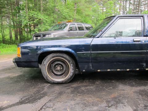 1991 Ford LTD Crown Victoria - A dark blue 1991 Ford Crown Victoria LTD is parked next to another vehicle under a tree line.