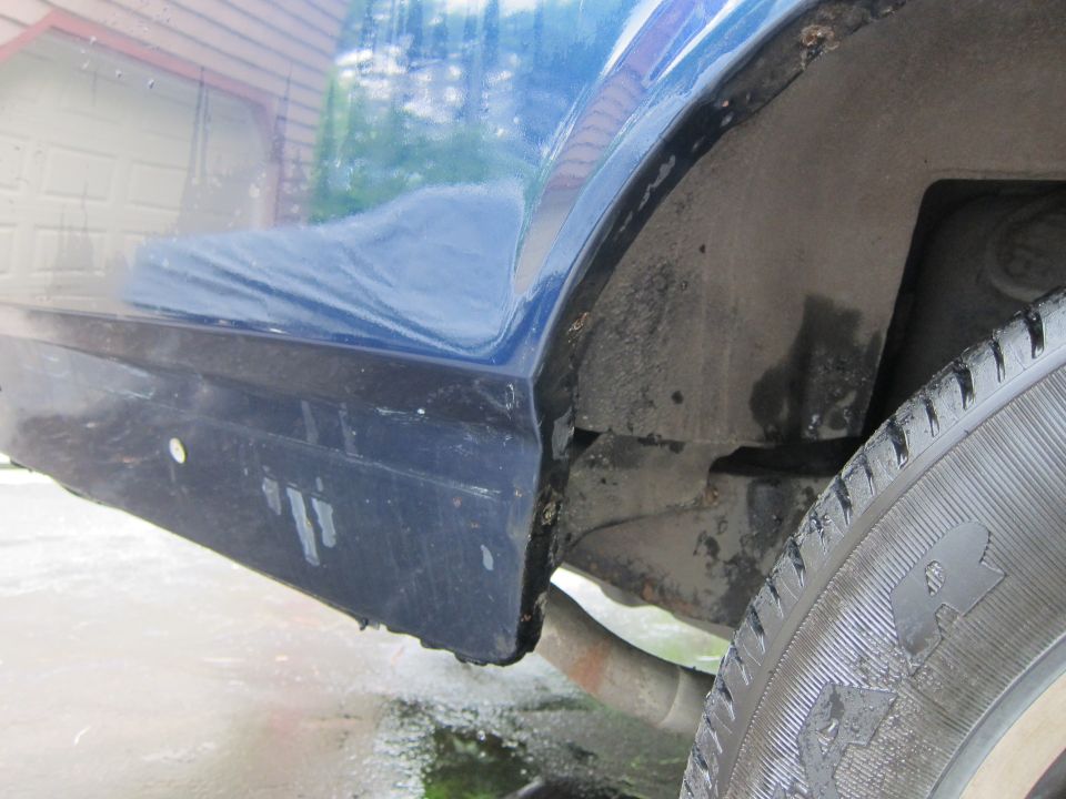 Close-up of a blue car's rear wheel well and lower body panel. Shows some rust and dirt
