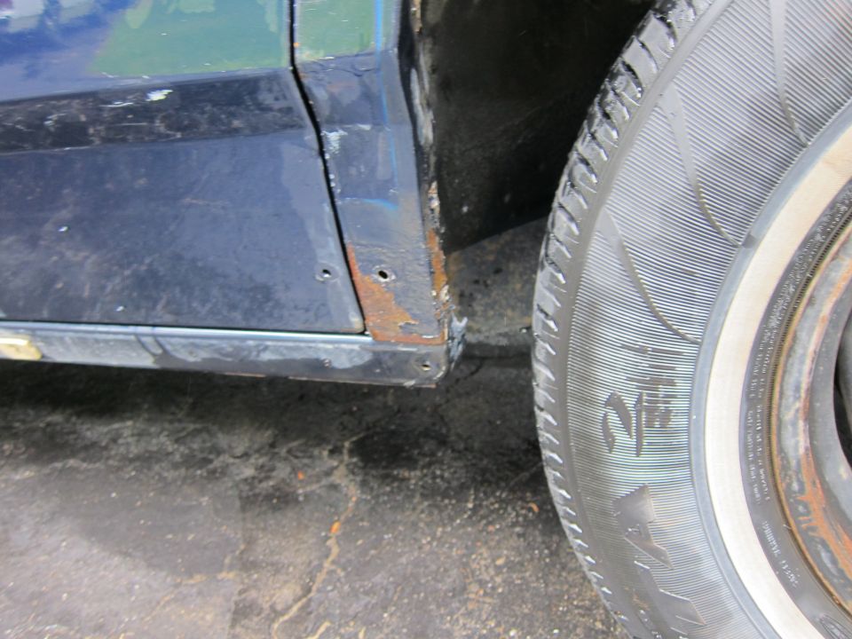 Dark blue car door with rust on the lower edge and fender seam, next to a tire with a white stripe