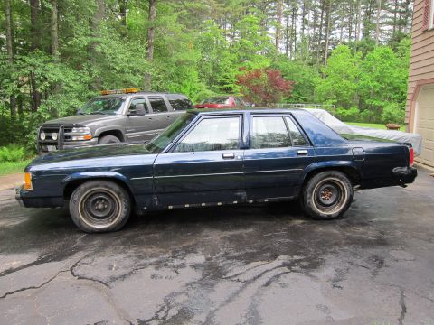1991 Ford LTD Crown Victoria - A dark blue sedan is parked in a driveway next to a gray SUV, with trees and a house in the background.