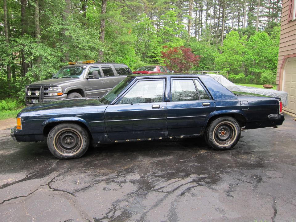 Dark blue sedan parked in driveway with a gray SUV behind it. Car has steel wheels and some rust on the lower body panels