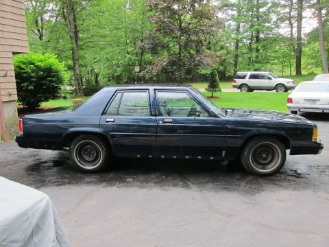 1991 Ford LTD Crown Victoria - A dark blue sedan is parked on a wet driveway next to a house, with other vehicles in the background.