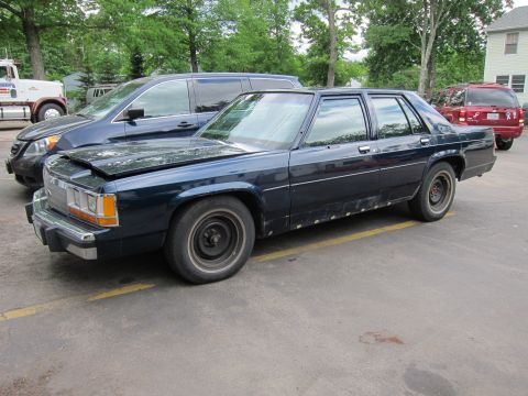 1991 Ford LTD Crown Victoria - A dark blue sedan with a damaged hood and visible rust on its lower body panels is parked in a lot.