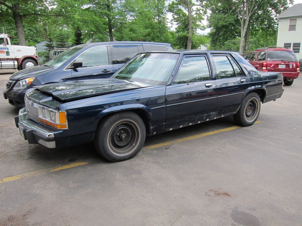 Dark blue sedan with dented hood and rust spots on lower body panels, parked on asphalt