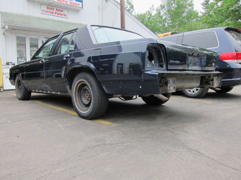 1991 Ford LTD Crown Victoria - A black sedan, likely a Ford Crown Victoria, is seen with significant rear damage, missing its bumper and taillight assembly.