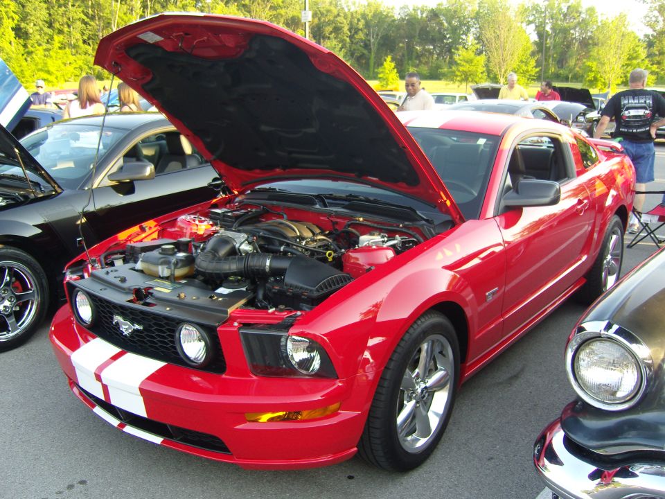 Red 2007 Ford Mustang with hood open, showing engine. White racing stripes on hood and front bumper. Another car is visible to the left