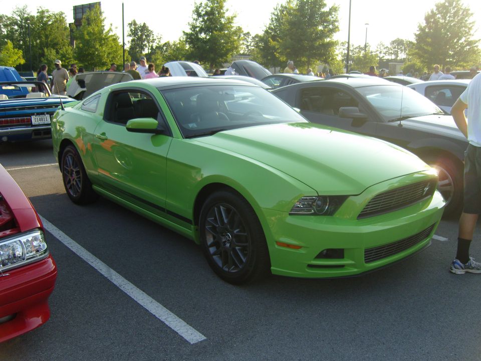 Green Ford Mustang parked in a lot, front 3/4 view. Black stripe on the side, dark wheels