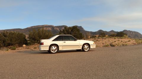 1989 Ford Mustang - A white coupe car is parked on a dirt road with mountains in the background under a clear blue sky.