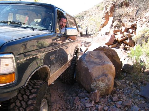 1994 Ford Bronco - A dark SUV with aggressive tires navigates a challenging rocky terrain, with another vehicle visible in the distance.