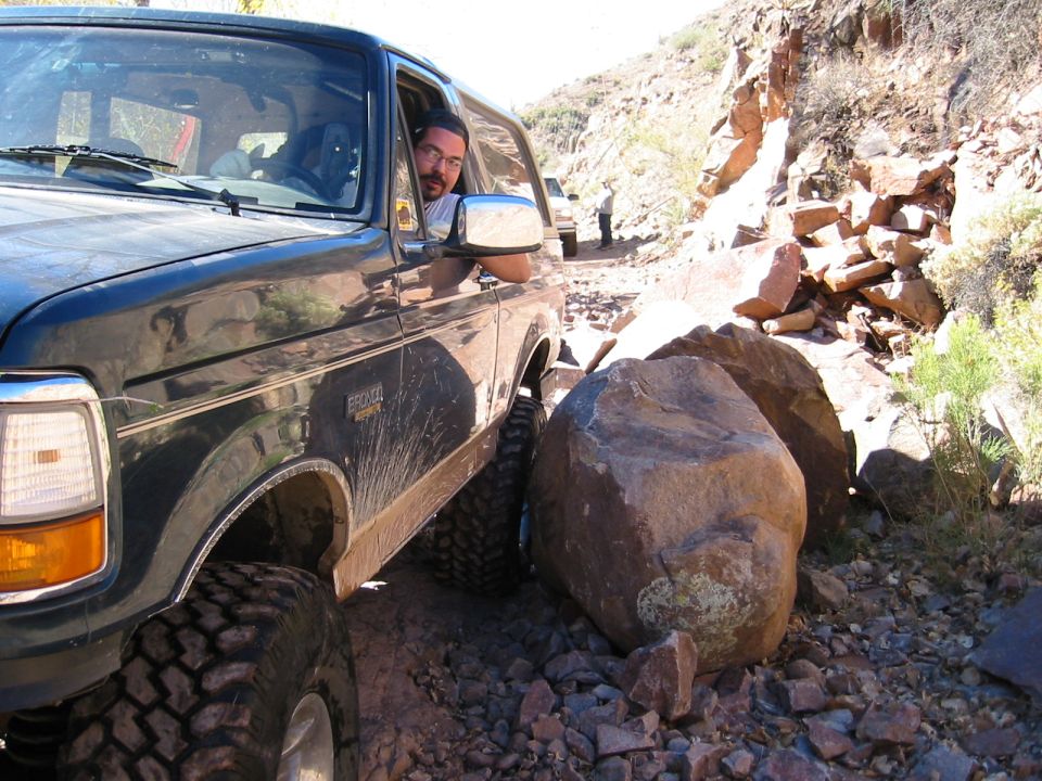 Dark green lifted Ford Bronco on mud-terrain tires easing past a boulder on a rocky desert trail