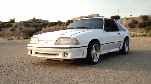 1989 Ford Mustang - A white Ford Mustang GT is parked on a gravel road with buildings in the background.