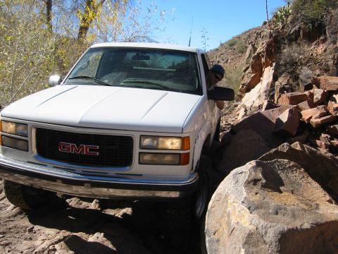 1994 Ford Bronco - A white GMC pickup truck maneuvers through a challenging, rocky off-road path under a clear blue sky.