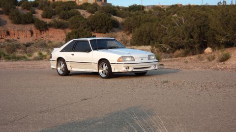 1989 Ford Mustang - A white 1989 Ford Mustang GT is parked on a gravel road with arid hills in the background.