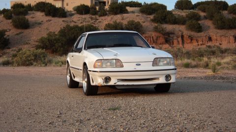 1989 Ford Mustang - A white Ford Mustang is parked on a dirt road with a house and arid landscape in the background.