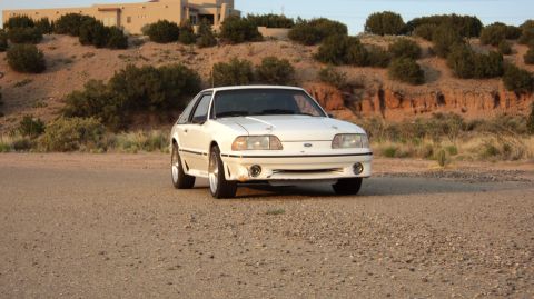 1989 Ford Mustang - A white 1989 Ford Mustang is parked on a dirt road with a building and arid landscape in the background.