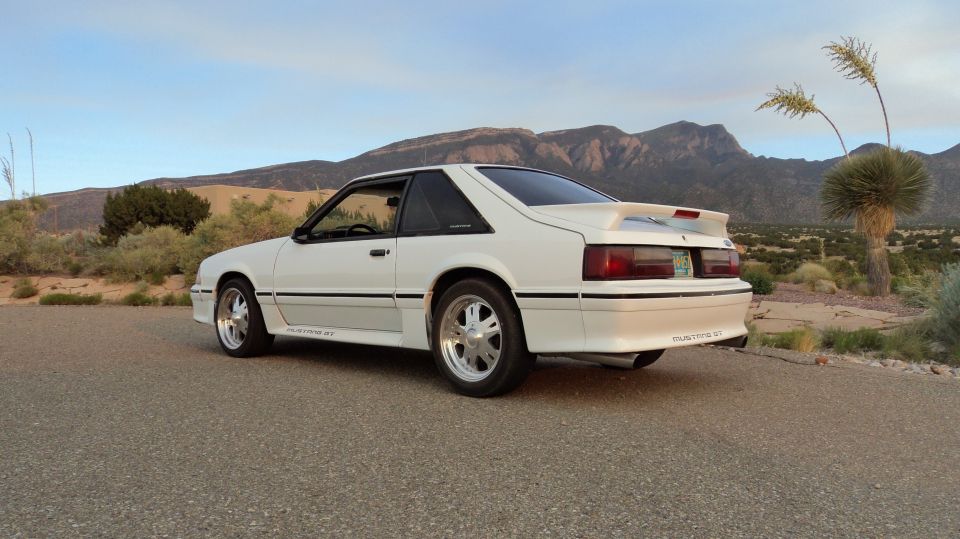 White 1987 Ford Mustang GT coupe parked on asphalt with desert mountains in background