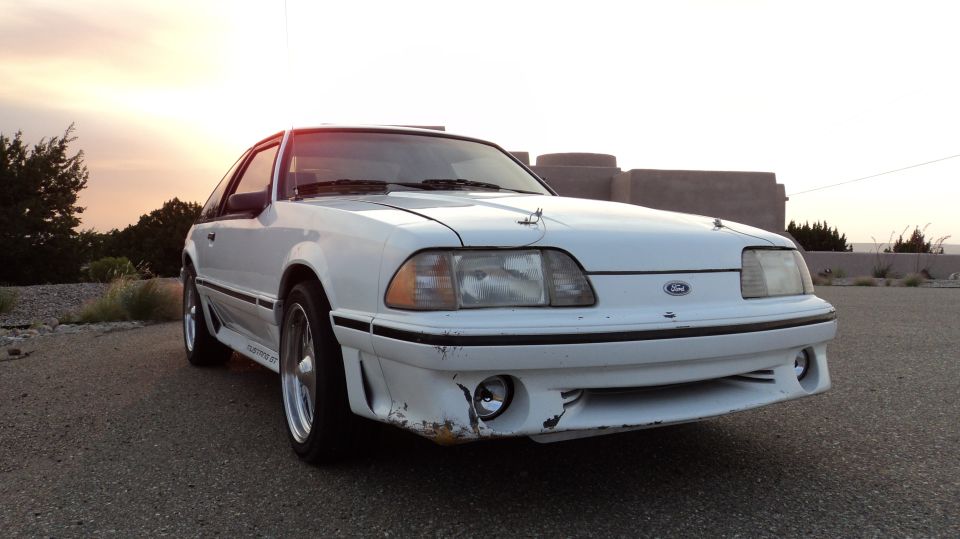 White Ford Mustang with aftermarket front bumper and hood pins, parked outside at sunset