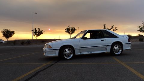 1989 Ford Mustang - A white Ford Mustang GT is parked in a lot as the sun sets in the background.