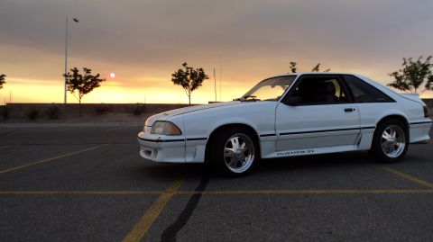 1989 Ford Mustang - A white 1989 Ford Mustang GT is parked in a lot during sunset, with the 'Mustang GT' badging visible on the side.