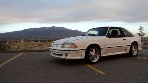 1989 Ford Mustang - A white 1989 Ford Mustang GT is parked in a lot, with mountains visible in the distance under a cloudy sky.