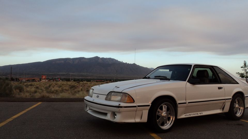 White 1989 Ford Mustang GT parked in a lot with mountains and cloudy sky