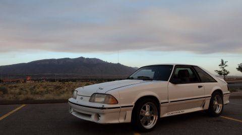 1989 Ford Mustang - A white Ford Mustang hatchback is parked outdoors with a mountain range visible in the distance.