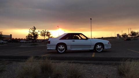 1989 Ford Mustang - A white 1989 Ford Mustang GT is parked in a lot as the sun sets behind it.