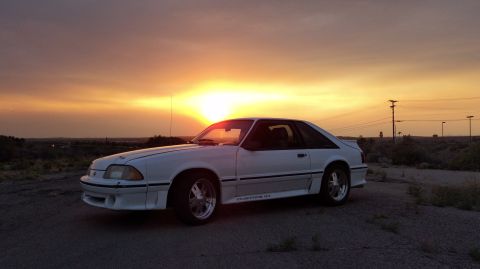 1989 Ford Mustang - A white 1987 Ford Mustang GT coupe is seen from a 3/4 front angle against a vibrant sunset sky.