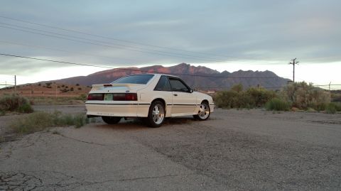 1989 Ford Mustang - A white coupe with a spoiler is parked on a gravel lot with mountains in the distance.