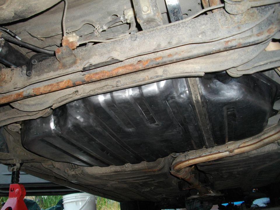 Underside view of a car's fuel tank and suspension components. Some rust is visible on the frame rails