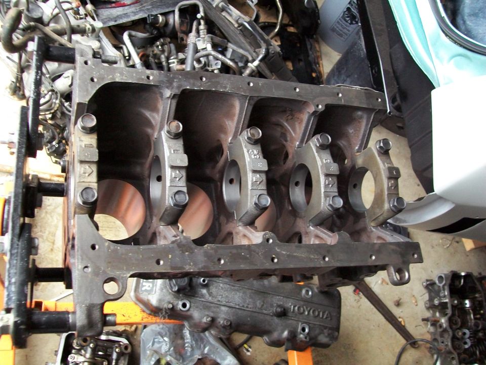Engine block on stand, with a Toyota valve cover visible below it. Some engine parts are in the background