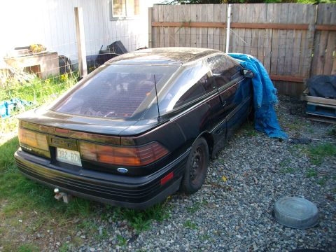 1989 Ford Probe - The rear of a black 1989 Ford Probe is visible, parked on a gravel lot with a blue tarp partially covering the vehicle.