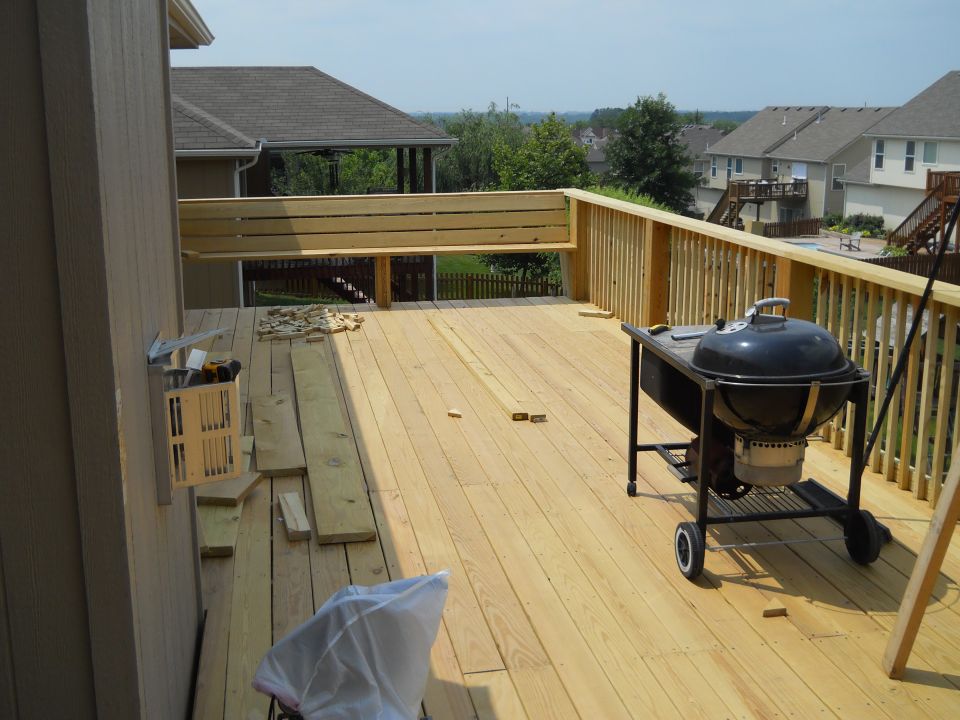 Wooden deck under construction with lumber, tools, and a black kettle grill. Houses and trees are visible in the background