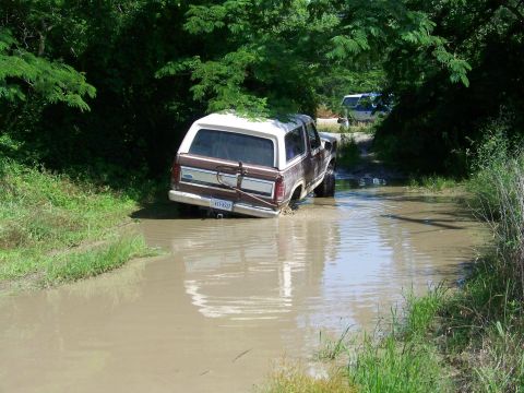1985 Ford Bronco - A brown and white Ford Bronco navigates a flooded dirt path, with another vehicle visible in the background.