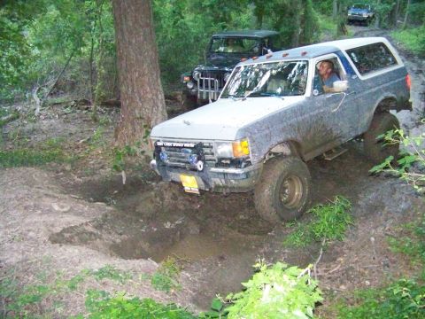 1985 Ford Bronco - Two off-road vehicles tackle a challenging, muddy path through a wooded area.